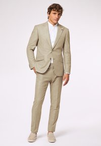Beige Leinenanzug mit maßgeschneidertem Schnitt, einreihiger Blazer und gerade geschnittenen Hosen. Darunter ein weißes Hemd mit Kragen, kombiniert mit hellen Mokassins.
