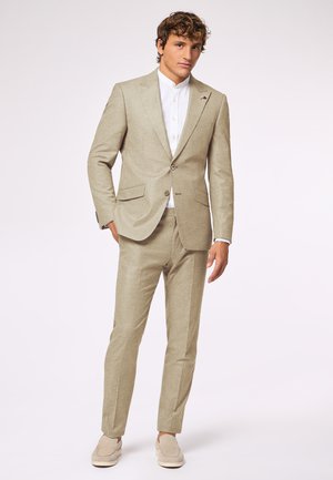 Beige Leinenanzug mit maßgeschneidertem Schnitt, einreihiger Blazer und gerade geschnittenen Hosen. Darunter ein weißes Hemd mit Kragen, kombiniert mit hellen Mokassins.