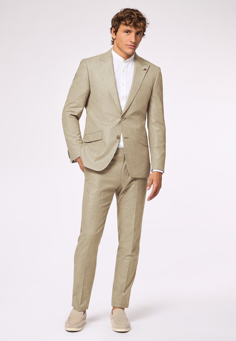 Beige Leinenanzug mit maßgeschneidertem Schnitt, einreihiger Blazer und gerade geschnittenen Hosen. Darunter ein weißes Hemd mit Kragen, kombiniert mit hellen Mokassins.