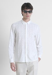 Camisa blanca de botones con un patrón geométrico texturizado, de manga larga y cuello puntiagudo. Combinada con pantalones de color claro y gafas de sol.