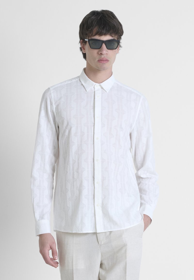 Camisa blanca de botones con un patrón geométrico texturizado, de manga larga y cuello puntiagudo. Combinada con pantalones de color claro y gafas de sol.