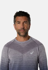 Långärmad sporttröja i gradientlila, med texturerade mönster och en logotyp på bröstet; tillverkad av lätt och andningsbart tyg.