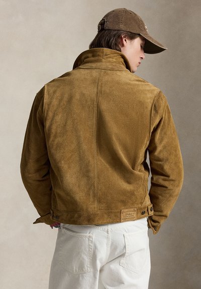 Νεαρός άνδρας που φοράει καφέ σαμουά μπουφάν και ταιριαστό καπέλο, εμφανιζόμενος από πίσω, με λευκό παντελόνι σε ουδέτερο φόντο.