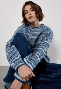 Young woman sitting with one knee up, wearing a blue patterned knitted sweater, dark blue jeans, and brown socks against a plain background.