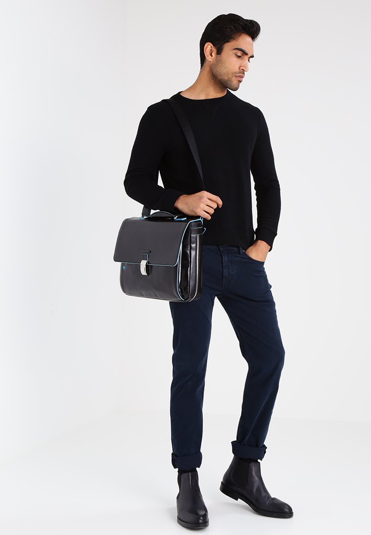 Borsa messenger in pelle nera con dettagli blu, chiusura a patta e hardware argentato, indossata da un modello con un outfit composto da un maglione nero e pantaloni blu navy.