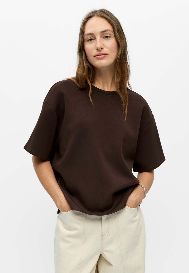 Mujer con cabello largo y castaño que lleva una camisa de manga corta suelta de color marrón oscuro y pantalones beige claros, manos en los bolsillos, fondo neutro.
