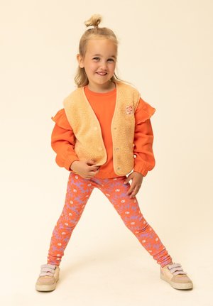 Oranje shirt met lange mouwen gecombineerd met een pluizig beige vest en legging met een bloemmotief in roze en lavendel. Schoenen met meerdere bandjes.