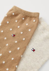 Bruine en crèmekleurige sokken; de ene met witte stippen, de andere effen. Beide hebben logo-accenten in marineblauw, rood en wit. Zachte gebreide textuur.