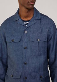 Giacca camicia in lino blu con colletto, due tasche sul petto e due tasche laterali. Presenta bottoni neri e un tessuto testurizzato.