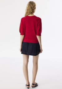 Czerwony sweter z krótkim rękawem, z prążkowanym dekoltem i dołem, w zestawie z ciemną spódnicą dżinsową z widocznymi szwami.