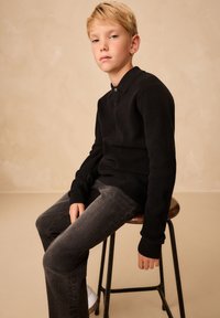 Pull polo noir en maille avec manches longues, doté d'un col et de détails à boutons, accompagné d'un jean gris foncé et de baskets blanches.