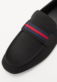 Sorte loafers laget av glatt materiale med polstret topp. Et blått og rødt stripet elastisk bånd over fronten gir detalj. Rund tådesign.