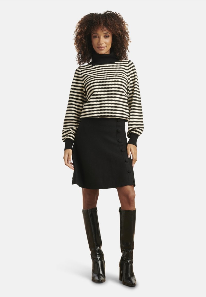 Gestreifter schwarze und weiße Pullover mit Rollkragen, kombiniert mit einem schwarzen Rock mit seitlichen Knöpfen. Das Model trägt knielange schwarze Stiefel.