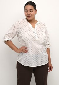 Blusa blanca de botones con un patrón de puntos negros, fabricada en un material suave y con un corte relajado. Presenta un escote sin cuello y mangas enrolladas de 3/4.