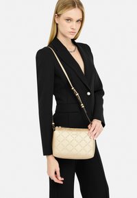 Borsa a tracolla beige con motivo trapuntato e dettagli del logo, dotata di una tracolla in catena color oro e chiusura a zip. Materiale sintetico liscio.