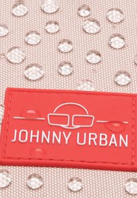 Beige szövet, tiszta vízcseppekkel, és egy piros gumicímke, amelynek fehér szövege "JOHNNY URBAN" egy szemüveg grafikája felett.