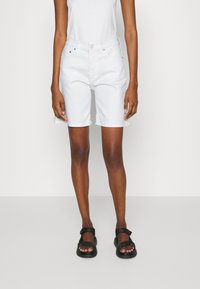 Persona con pantalones cortos blancos hasta la rodilla, una camiseta sin mangas blanca y sandalias negras de punta abierta, de pie contra un fondo claro liso.