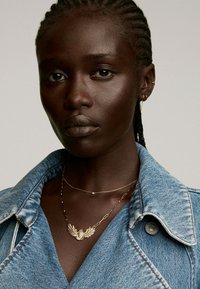 Chaqueta de mezclilla con cuello, de color azul claro y tela texturizada. La joyería incluye cadenas de oro en capas con un colgante de alas.