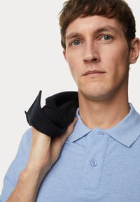 Helles blaues Polohemd mit strukturiertem Finish, ausgestattet mit einem Kragen und einem Knopf. Ein schwarzes Kleidungsstück ist lässig über die Schulter drapiert.