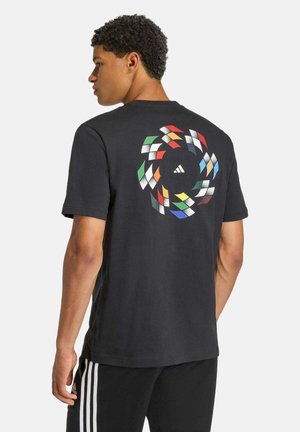 Jeune homme portant un t-shirt noir avec un motif circulaire coloré et le logo Adidas dans le dos, associé à un pantalon noir à rayures blanches.