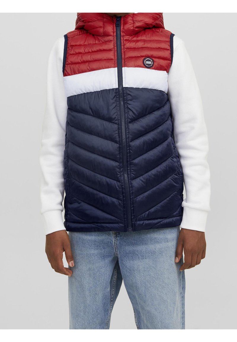 Person som bär en ärmlös, huva puffer väst med röda, vita och marinblå färgblock över en vit långärmad tröja och ljusblå jeans.