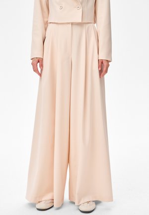 Kobieta ubrana w beżowe spodnie z wysokim stanem i szerokimi nogawkami, zestawione z dopasowaną krótką marynarką i beżowymi butami.