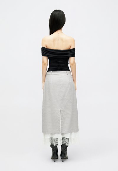 Moteris su ilgais juodais plaukais, vilkinti juodą palaidinę be petnešėlių, pilką midi sijoną su nugaroje įpjova, baltu nėriniuotu pamušalu ir juodais aukštakulniais batais, stovinti.