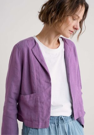 Linen, cropped jacket in light purple with a shawl collar, two front pockets, and a relaxed fit over a white T-shirt and blue pants.