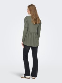 Vrouw met lang blond haar, gekleed in een olijfgroene top met lange mouwen, zwarte uitlopende broek en witte sneakers, staand en naar links kijkend.