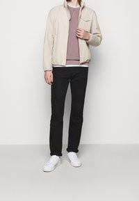 Giacca beige chiaro con zip e una tasca sul petto, indossata sopra un dolcevita malva e una camicia bianca, abbinata a pantaloni neri e sneakers bianche.