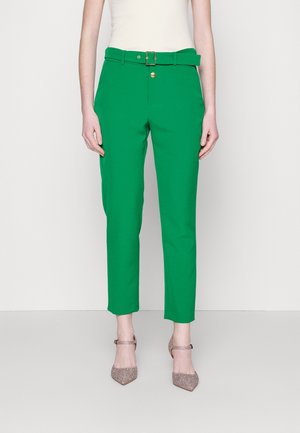 Woman wearing high-waisted green trousers with a matching belt and gold buckle, paired with sparkly pink pointed heels.