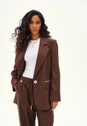 Kobieta o kręconych włosach ubrana w brązowy garnitur w prążki z różowymi akcentami, białą koszulę i warstwowe złote naszyjniki, stojąca z jedną ręką w kieszeni.