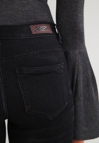 Jeans de mezclilla negra con una etiqueta de cuero. Presentan bolsillos traseros y una cintura ajustada, combinados con una camiseta de manga larga gris.