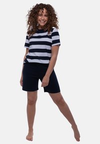 Hymyilevä nainen, jolla on kiharat hiukset, pukeutuneena laivastonsiniseen ja valkoiseen raidalliseen paitaan sekä laivastonsinisiin shortsit, seisoo paljain jaloin rentoutuneessa asennossa.