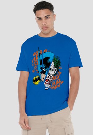 Man wearing a blue T-shirt with a split graphic of Batman and Joker faces and repeated orange "HA" text background.