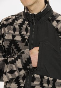 Fleecejacke mit einem grauen und schwarzen geometrischen Muster, einem einfarbigen schwarzen Reißverschluss vorne und einer Brusttasche. Weicher Stoff, hoher Kragen.