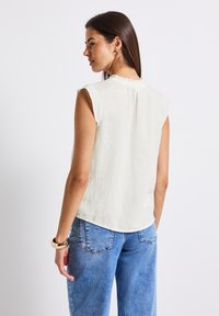 Elfenbensfärgad ärmlös blus med volangdetaljer vid halsen och slät textur, tillsammans med ljusblå, avslappnade jeans med bakfickor.