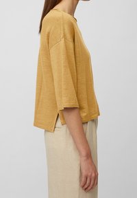 Femme portant un haut tricoté moutarde à manches trois quarts avec des fentes sur les côtés, et un pantalon beige clair taille haute, se tenant de profil devant un fond gris.