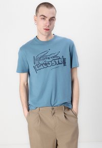 Jasnoniebieski bawełniany T-shirt z ciemną grafiką krokodyla i napisem "Lacoste". Krótkie rękawy, okrągły dekolt i luźny krój.