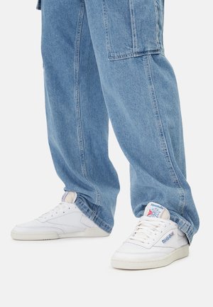 Białe trampki z teksturowanym cholewką ze skóry, ozdobione niebieskimi akcentami logo Reebok, zestawione z luźnymi, jasnoniebieskimi dżinsami.