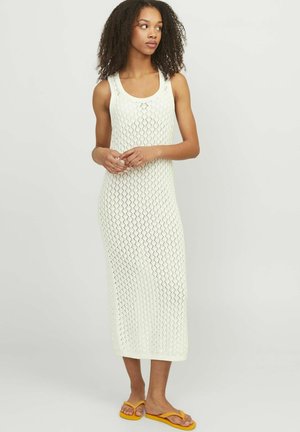Vestido midi blanco de punto con un patrón de diamantes, escote redondeado y tirantes anchos. Combinado con chanclas amarillas, creando un look informal.
