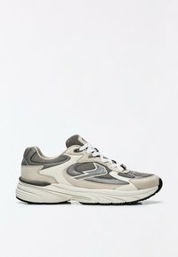 Chaussure de sport grise et beige avec lacets blancs et tissu en mesh, présentée sur fond blanc, vue de côté.