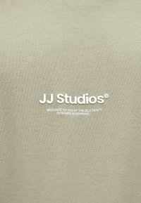 Jasnooliwkowa koszulka o miękkiej, gładkiej fakturze. Na froncie biały napis: "JJ Studios" oraz dodatkowe szczegóły brandingowe poniżej.