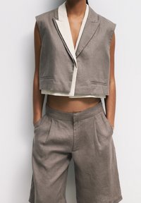 Persona con un blazer cortado sin mangas en color topo sobre una blusa blanca y pantalones cortos de talle alto plisados a juego, de pie con las manos en los bolsillos.