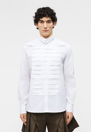 Homme aux cheveux bouclés portant une chemise blanche à boutons avec plusieurs bandes horizontales en tissu sur la poitrine et un pantalon marron à motifs.