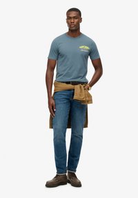 Camiseta azul de manga corta con estampado verde, combinada con jeans de mezclilla y botas marrones con cordones. Hay una camisa color beige atada a la cintura.