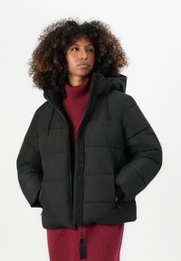 Jachetă puffer neagră cu glugă, având textură matlasată, buzunare laterale și fermoar frontal. Purtați-o peste un pulover tricotat bordeaux cu guler înalt.