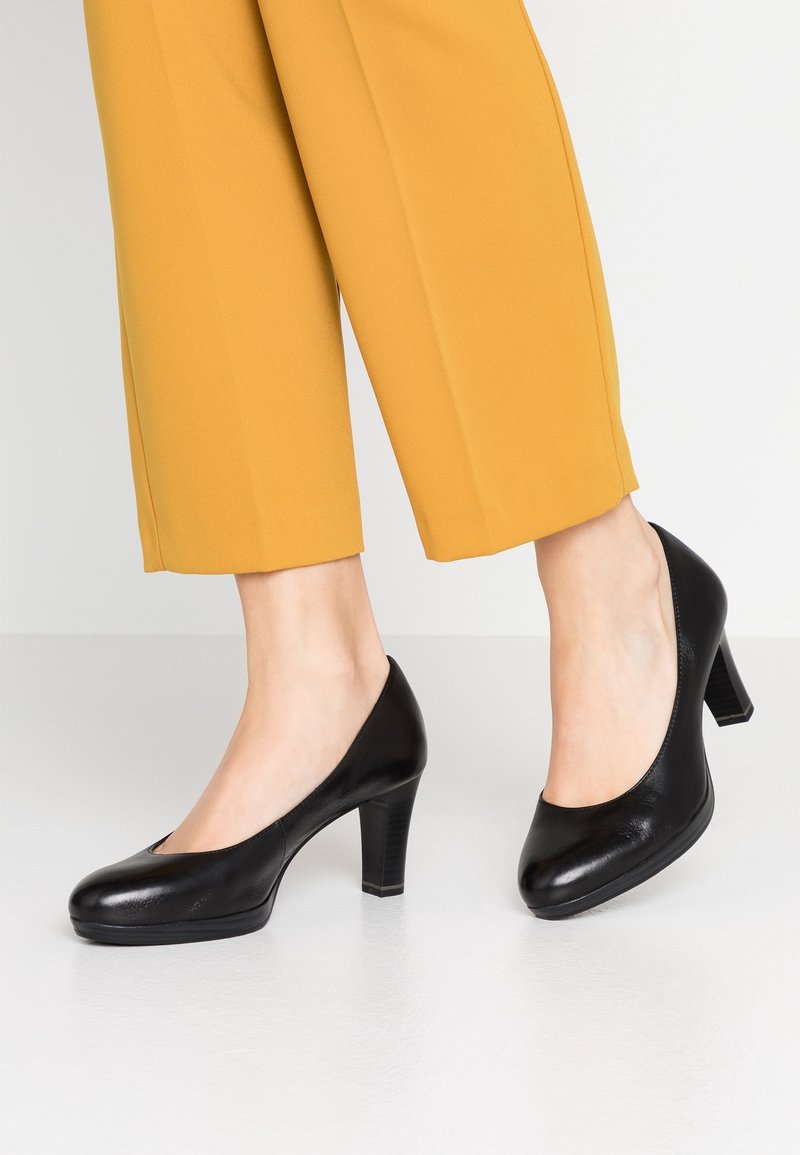 Personne portant un pantalon court jaune moutarde et des escarpins noirs en cuir à talon moyen, debout sur un sol gris clair.