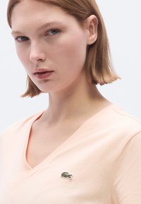 Ροδακινί T-shirt με V-λαιμόκοψη από μαλακό βαμβάκι, με μικρό πράσινο λογότυπο Lacoste στην αριστερή πλευρά του στήθους. Ομαλή υφή, casual σχεδίαση.