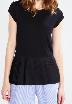 Vrouw draagt een zwart kortärmelig topje met geplooide zoom en lichtblauwe gestreepte broek, staand tegen een effen achtergrond.